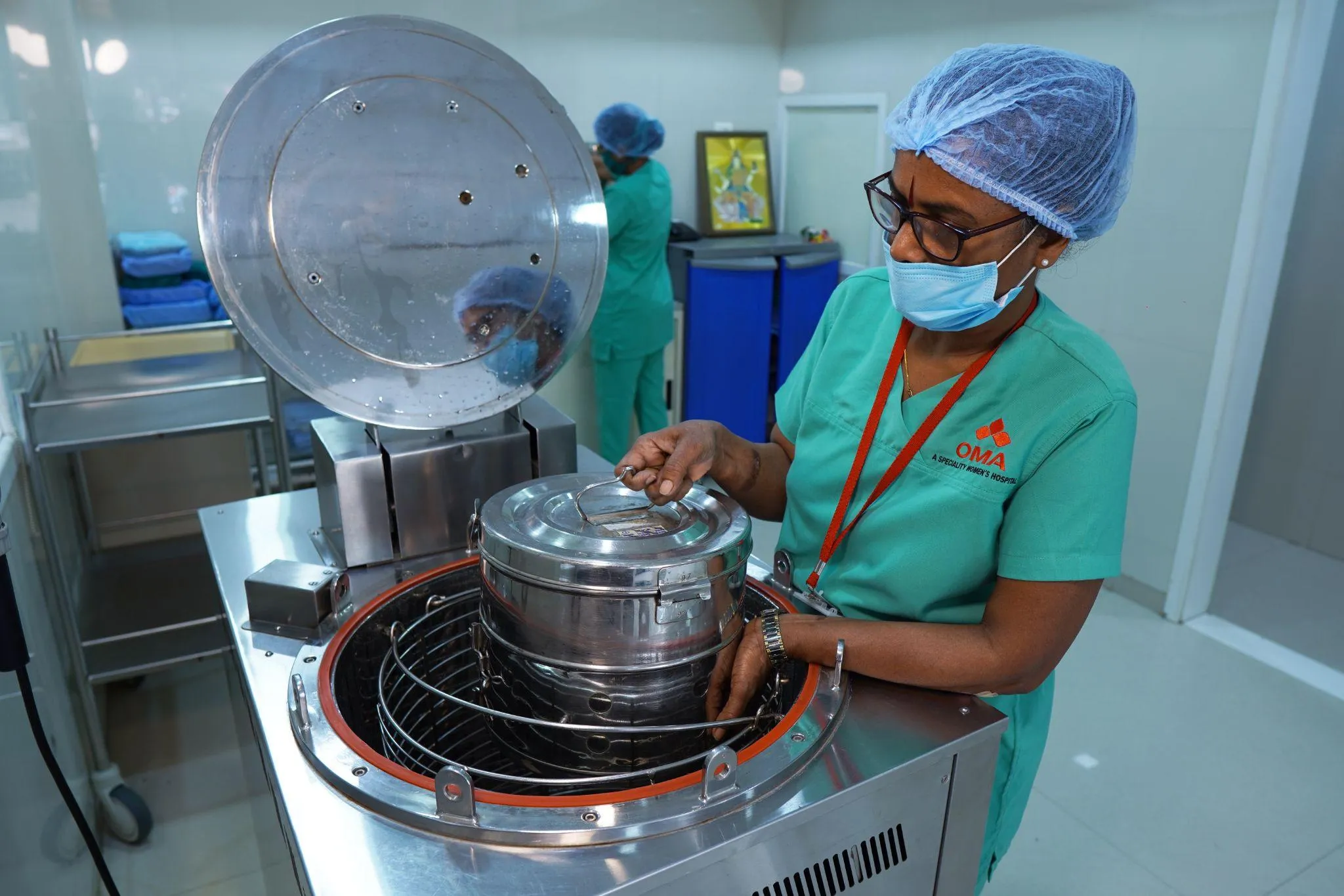 Female healthcare worker in scrubs and a blue cap uses an autoclave sterilizer in a hospital setting.