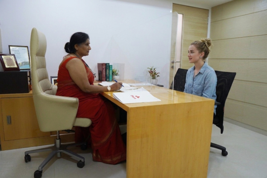 Two women sit across a wooden desk in an office; one in a red sari explains documents to a woman in a striped shirt wearing a face mask.