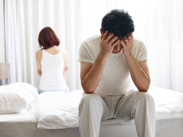 Picture1(80) Man sits on a bed with his head in his hands, looking stressed, while a woman sits apart with her back to him in a bright bedroom.