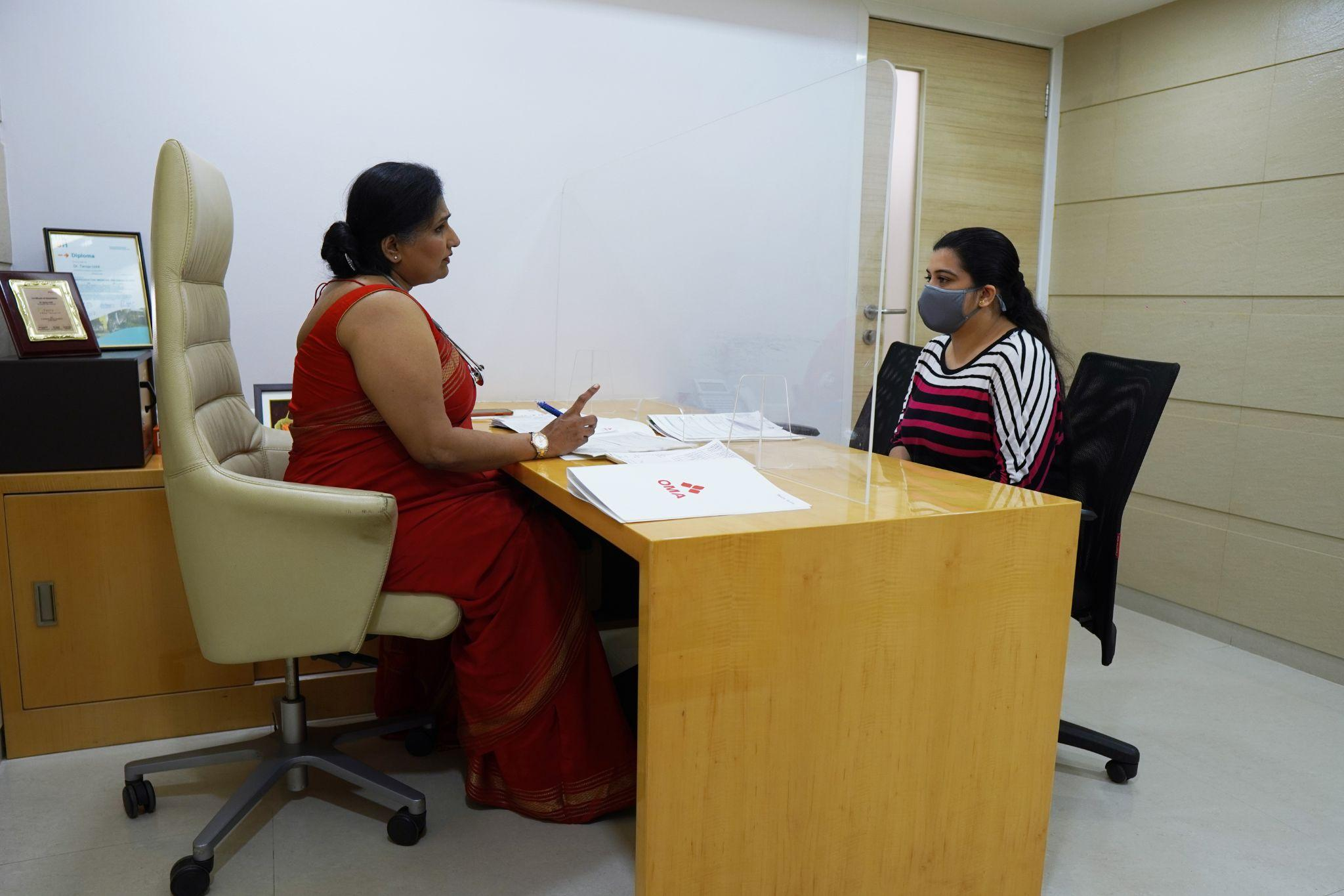 Two women sit across a desk with a clear partition between them; one in a red sari and the other in a striped top wearing a gray mask, discussing documents in an office.