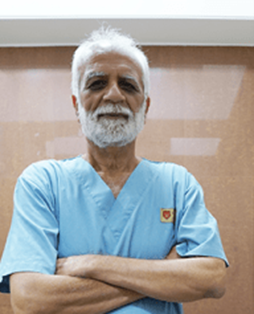 Older man with gray beard in blue medical scrubs, arms crossed, standing in a clinic or medical setting with a badge on his chest.