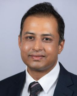 Professional headshot of a man in a dark suit and striped tie, smiling against a light gray background.