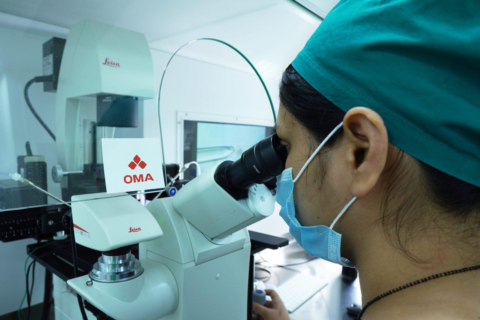 A lab technician looking into a Leica microscope, wearing a teal scrubs cap and blue mask in a cleanroom.