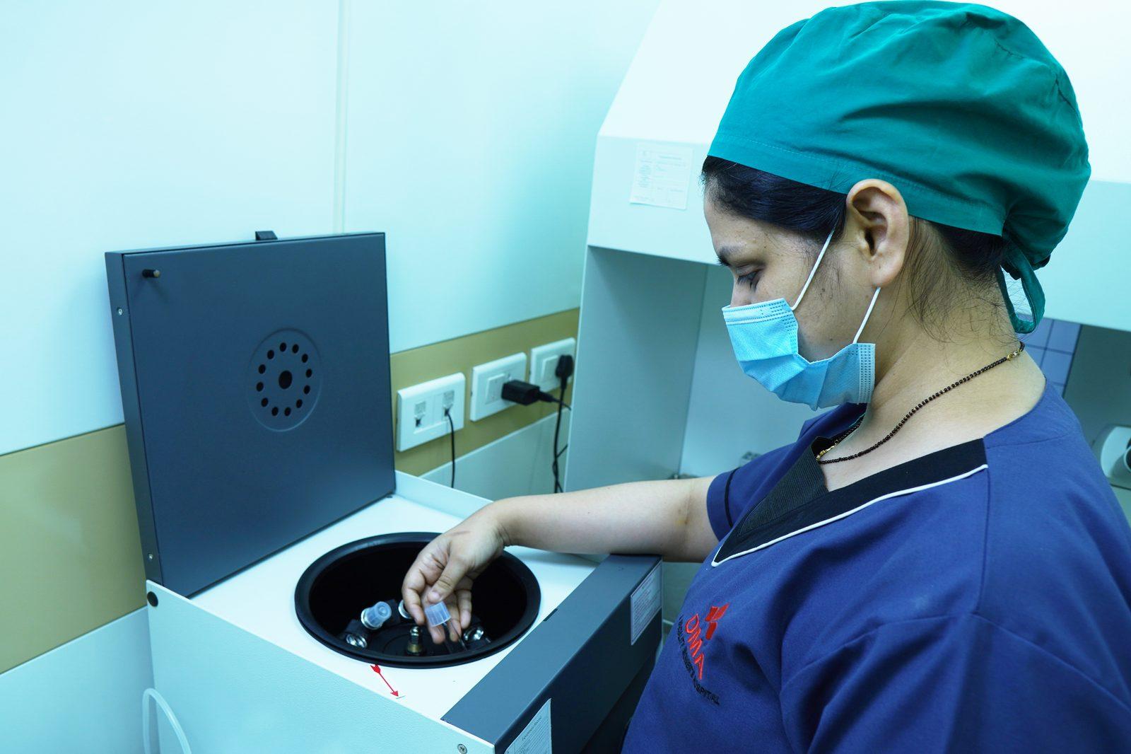 Nurse in scrubs and mask operates a laboratory centrifuge, placing tubes into the rotor for processing.