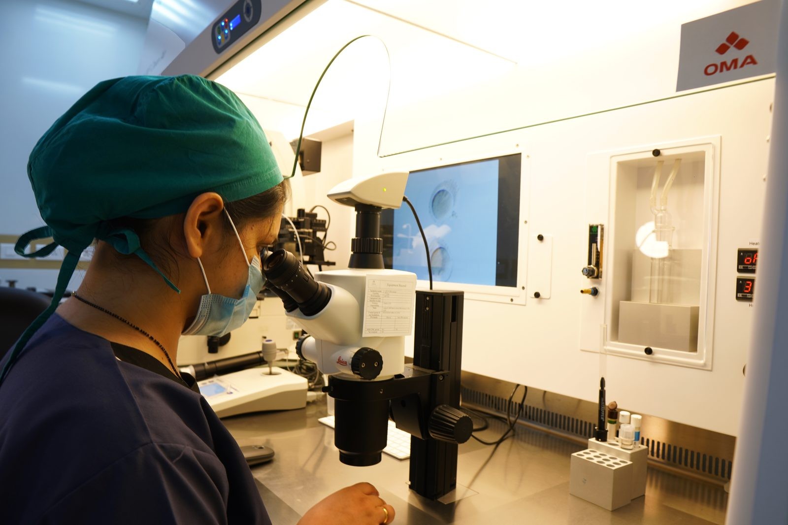 Female scientist in a green surgical cap and mask uses a Leica microscope at a clean lab bench.