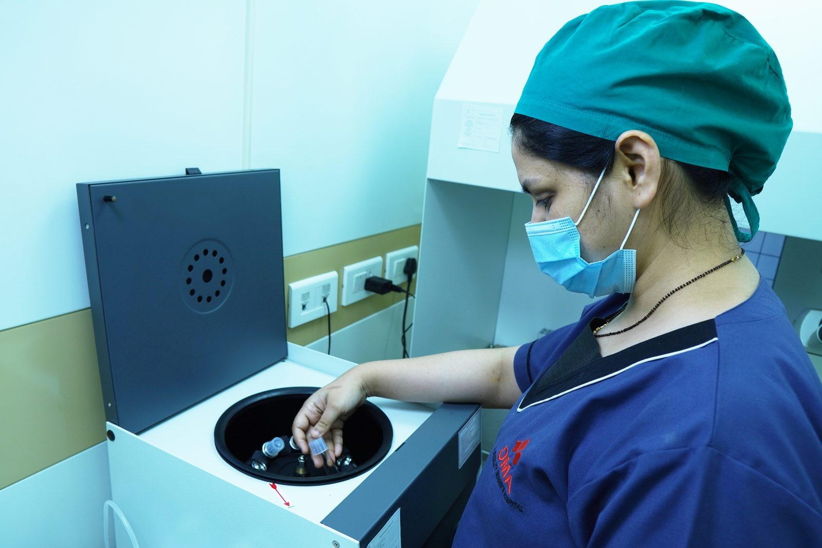 Healthcare worker in scrubs and mask loads tubes into an open centrifuge in a laboratory setting