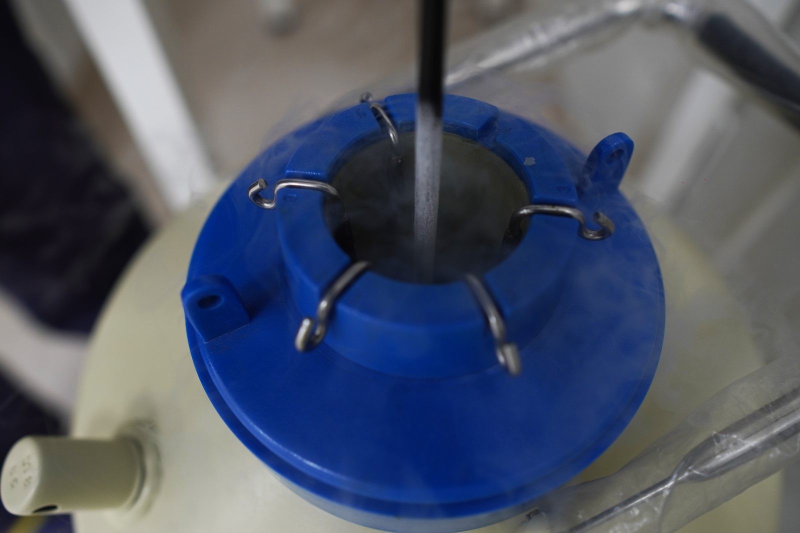 Close-up of a blue lab bottle lid with metal locking clips around an open center, a metal rod inserted downward into the opening.