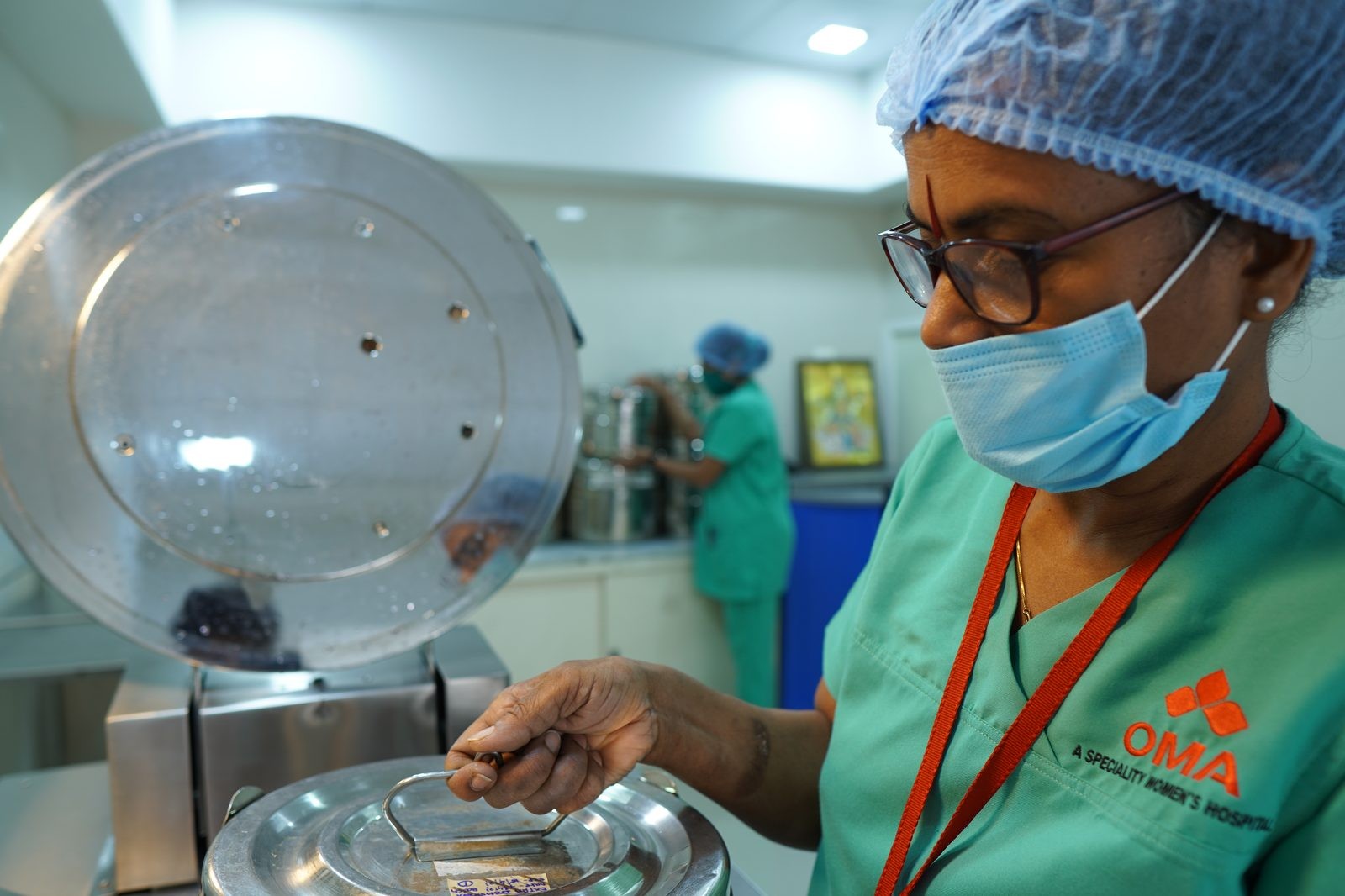 Nurse wearing a surgical cap and mask loads a sterile metal container into an autoclave in a hospital sterilization area.