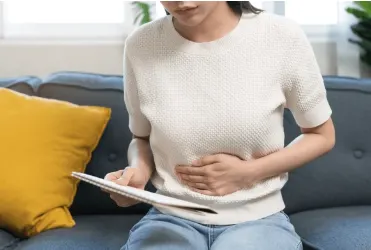 Woman on a sofa holds her abdomen and looks at a document, indicating stomach pain or discomfort in a home setting.
